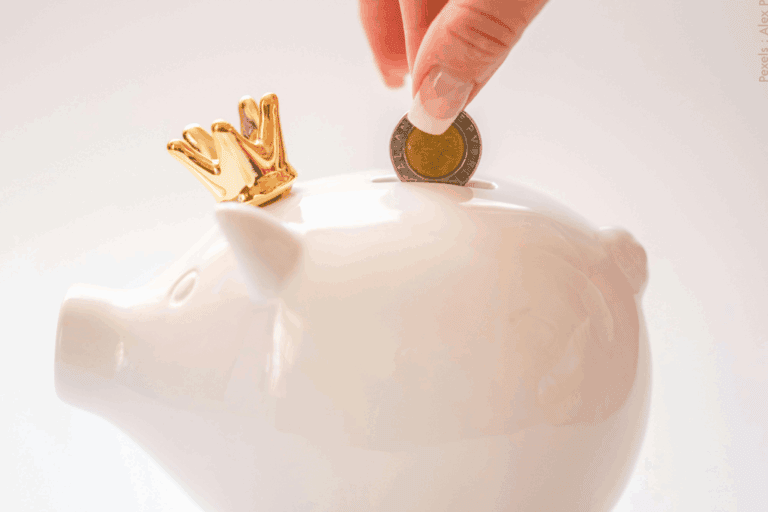 Doigts plaçant une pièce de monnaie dans une tirelire blanche en forme de petit cochon avec une couronne dorée sur la tête, symbolisant l'épargne et la finance.