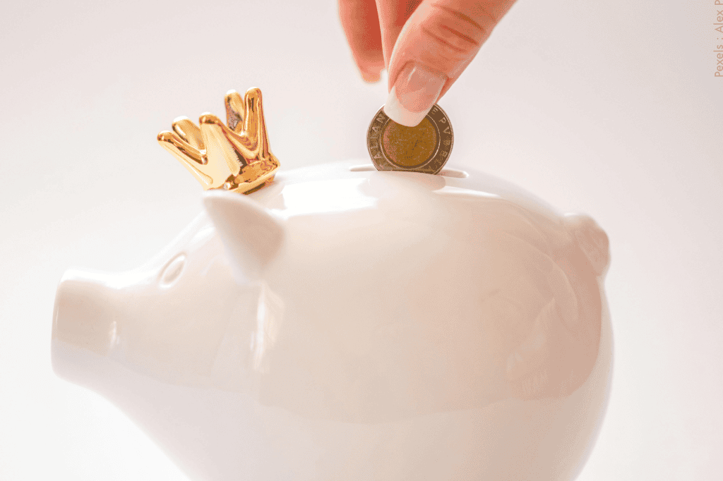Doigts plaçant une pièce de monnaie dans une tirelire blanche en forme de petit cochon avec une couronne dorée sur la tête, symbolisant l'épargne et la finance.